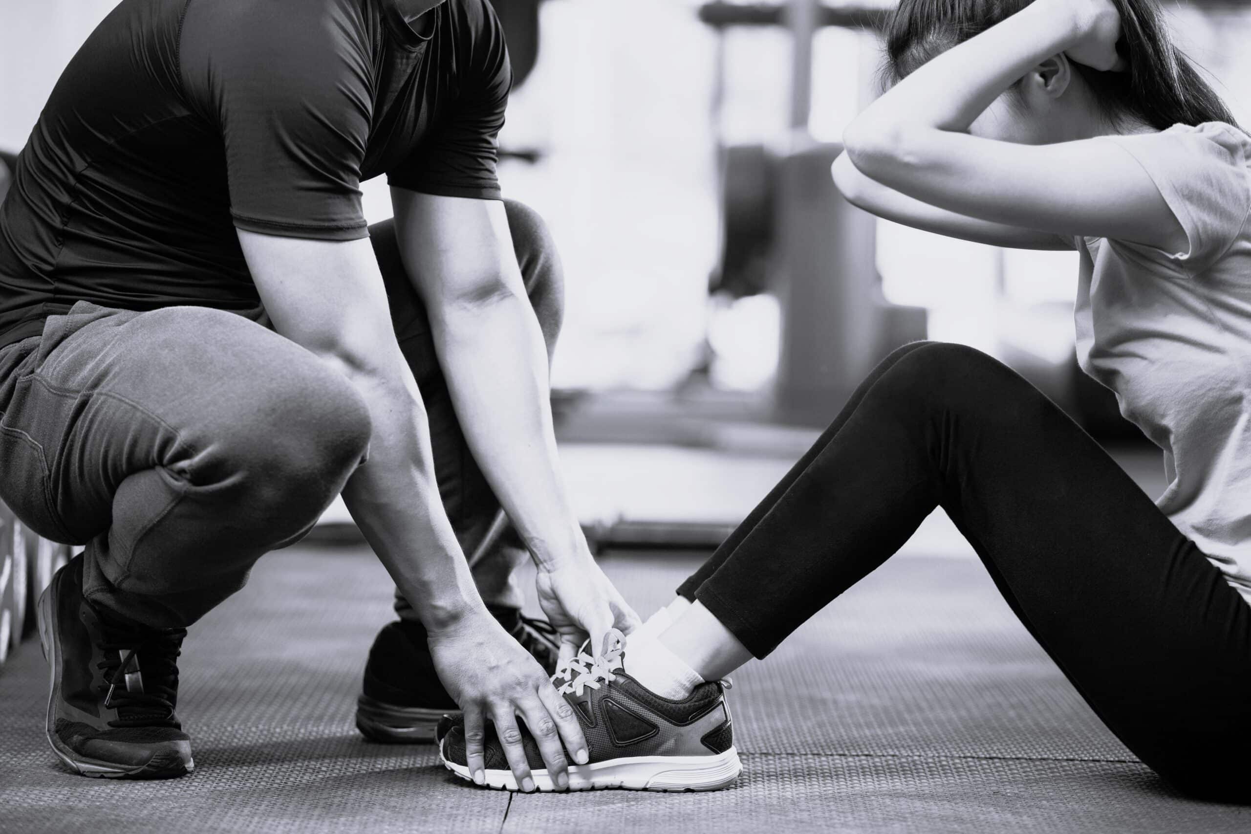 Entraîneur personnel aidant une femme à réaliser des abdominaux dans un gym, maintenant fermement ses chevilles sur le tapis de sol.