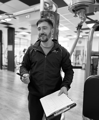 Coach Sportif en Salle de Musculation avec Plan d'Entraînement Entraîneur sportif souriant en veste noire dans une salle de sport, tenant un stylo et un presse-papiers avec des notes.