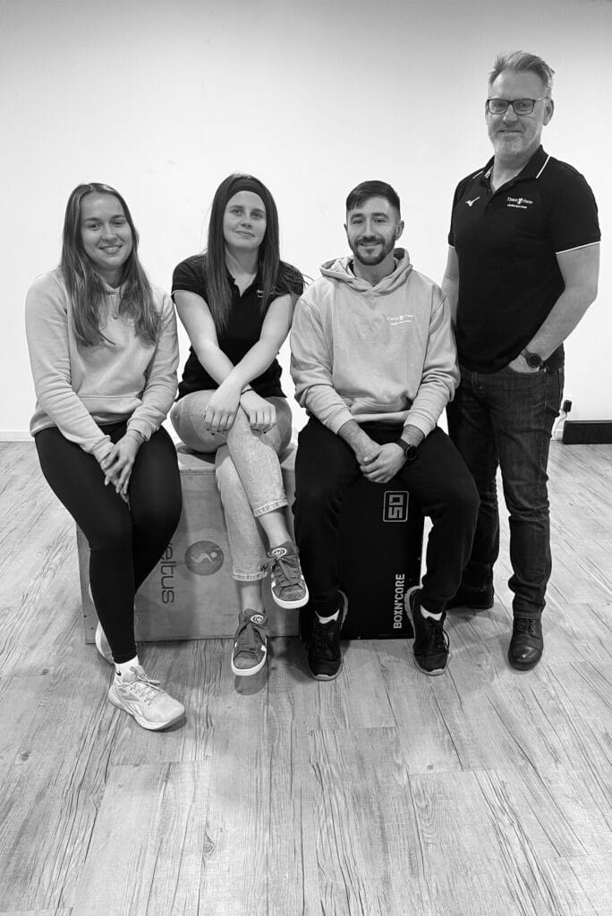 Équipe de sport souriante posant avec équipement de fitness Quatre personnes souriantes posent dans un gymnase; deux sont assises sur des boîtes de plyométrie 'eltus' et 'BOXN'CORE'.