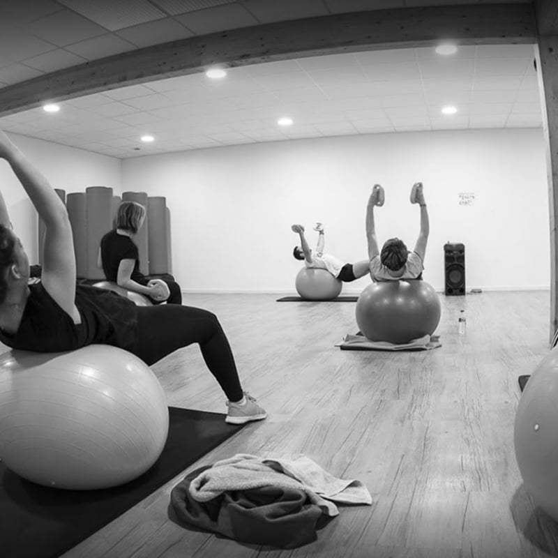 Exercices de gainage avec ballons de stabilité (Swiss balls) en noir et blanc, quatre personnes s'entraînent dans une salle lumineuse.