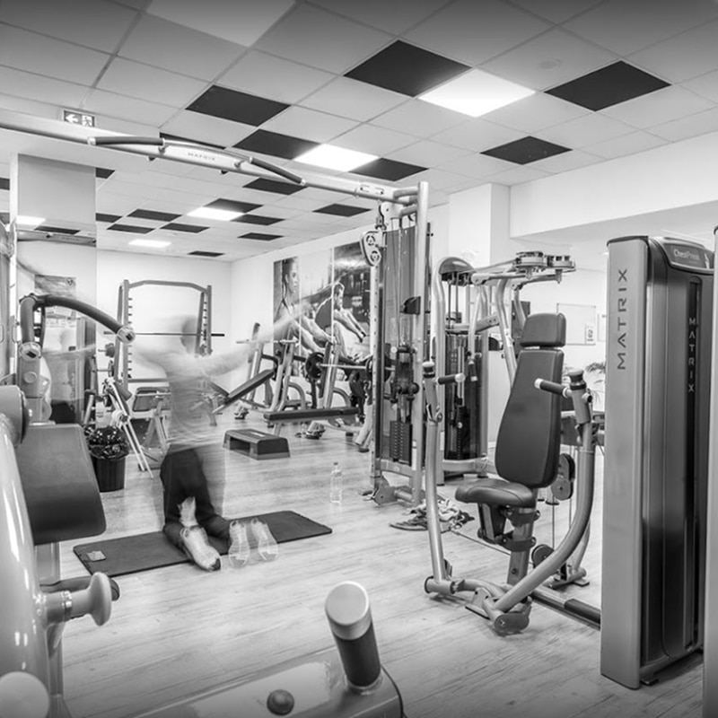 Scène intérieure de salle de sport en noir et blanc avec équipement MATRIX, une personne en mouvement flou faisant un exercice au sol, et des affiches murales.